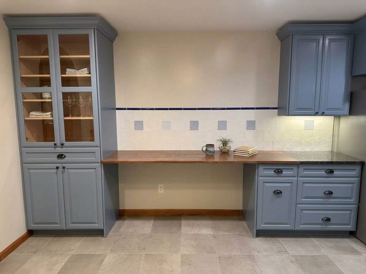 Built-in desk nook with tall glass-front cabinet and walnut countertop in a slate blue kitchen